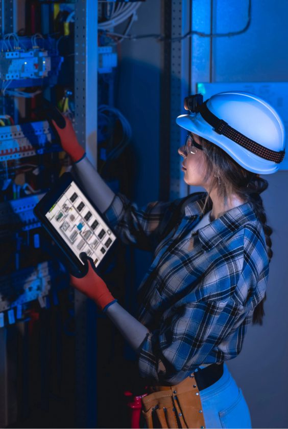 Eine Elektrikerin inspiziert einen elektrischen Schaltschrank im Serverraum und nutzt ein Tablet zur Diagnose und Kontrolle.